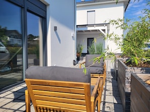 Un banco de madera y una silla con cojín en un patio empedrado con vistas a una casa moderna y un jardín.