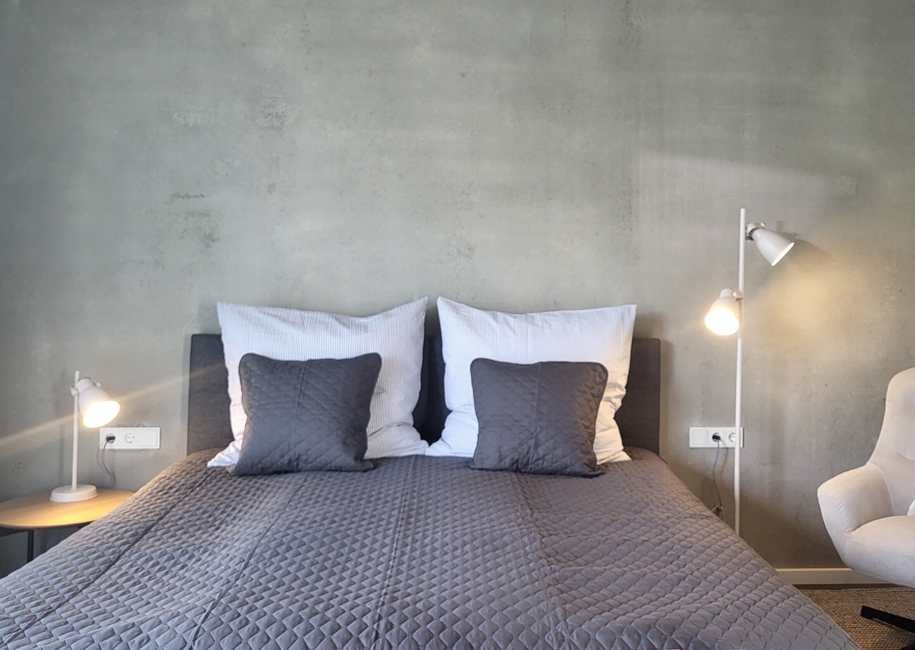 Una cama con un edredón gris, dos almohadas, una lámpara blanca a cada lado y una silla blanca.