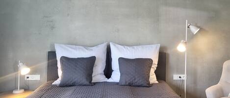 Una cama con un edredón gris, dos almohadas, una lámpara blanca a cada lado y una silla blanca.