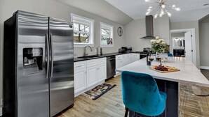 Una cocina moderna con refrigerador de acero inoxidable, armarios blancos, una isla central con una silla azul y una araña.