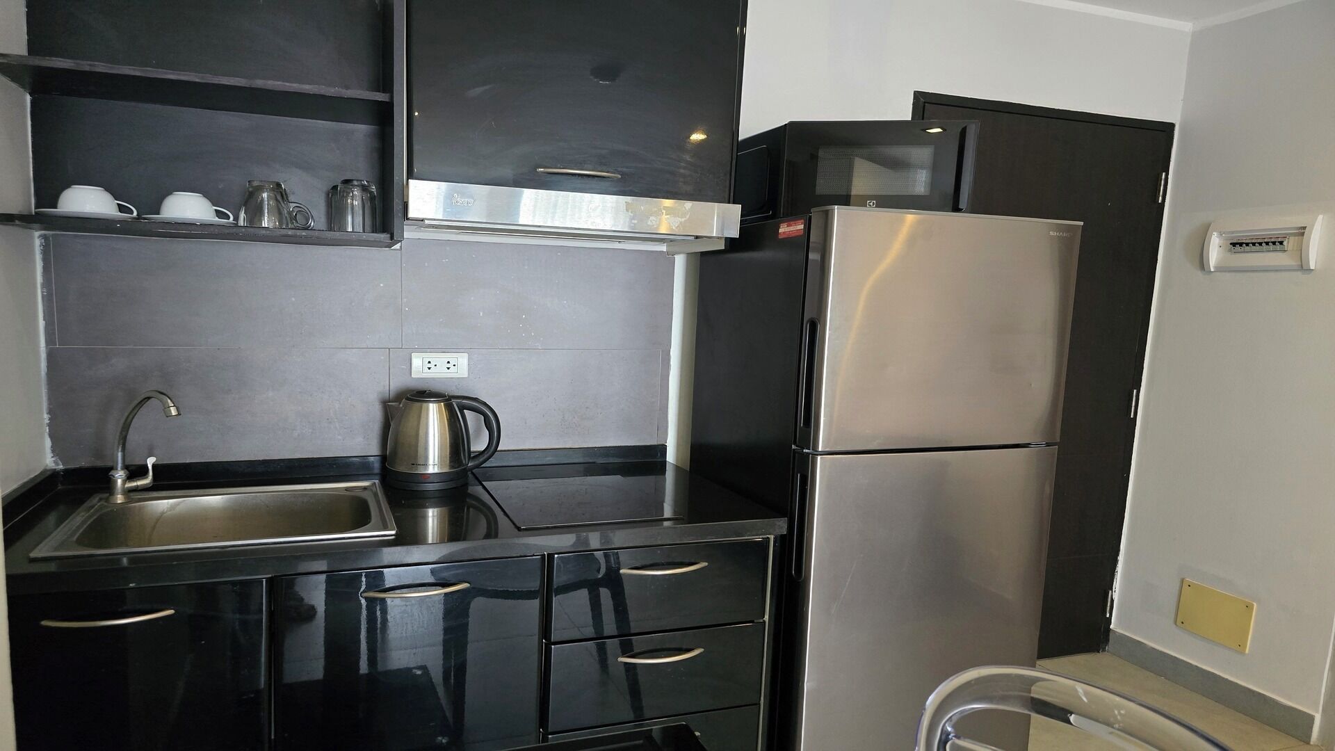 A compact kitchen with a stainless steel refrigerator, a kettle, and a sink with a faucet.