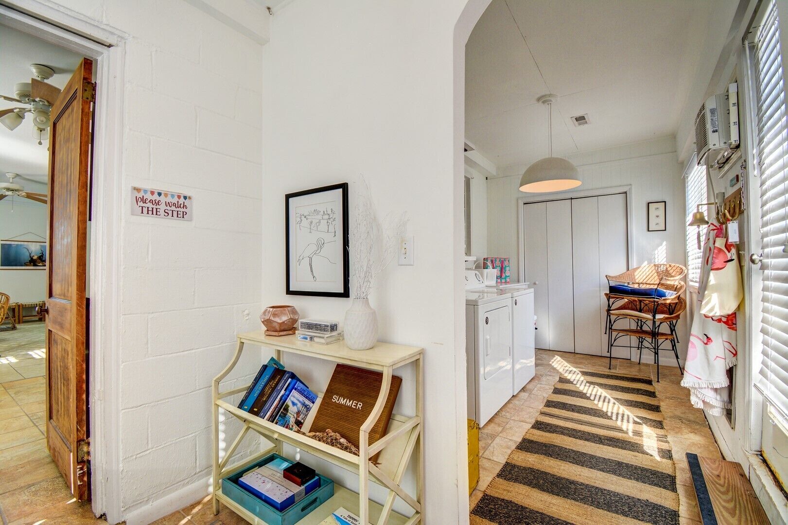 Een gang met een witte muur, een lijst met foto, een klein tafeltje met boeken, een wasmachine en een stoel.