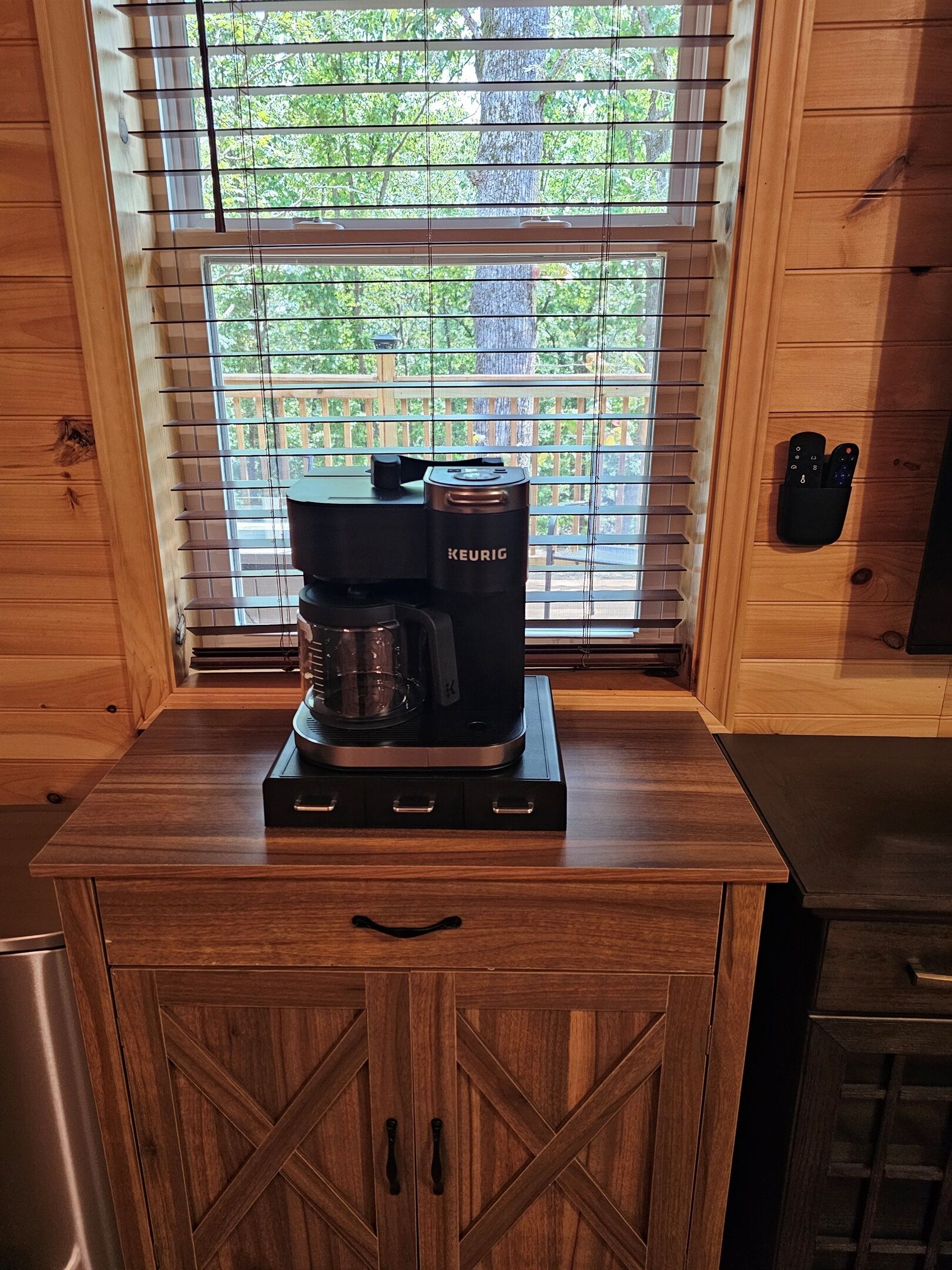 Une cuisine avec une machine à café Keurig posée sur un comptoir en bois, une fenêtre avec des stores et un meuble en bois doté de tiroirs.
