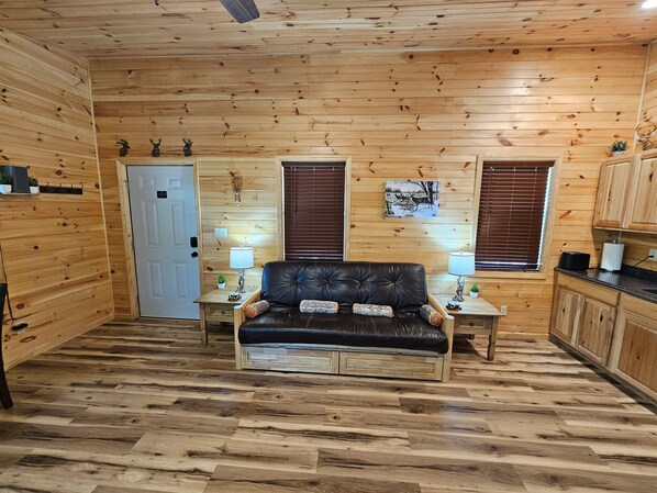 A cozy cabin interior with a leather sofa, wooden walls, a door, two windows with blinds, and a small kitchen area.