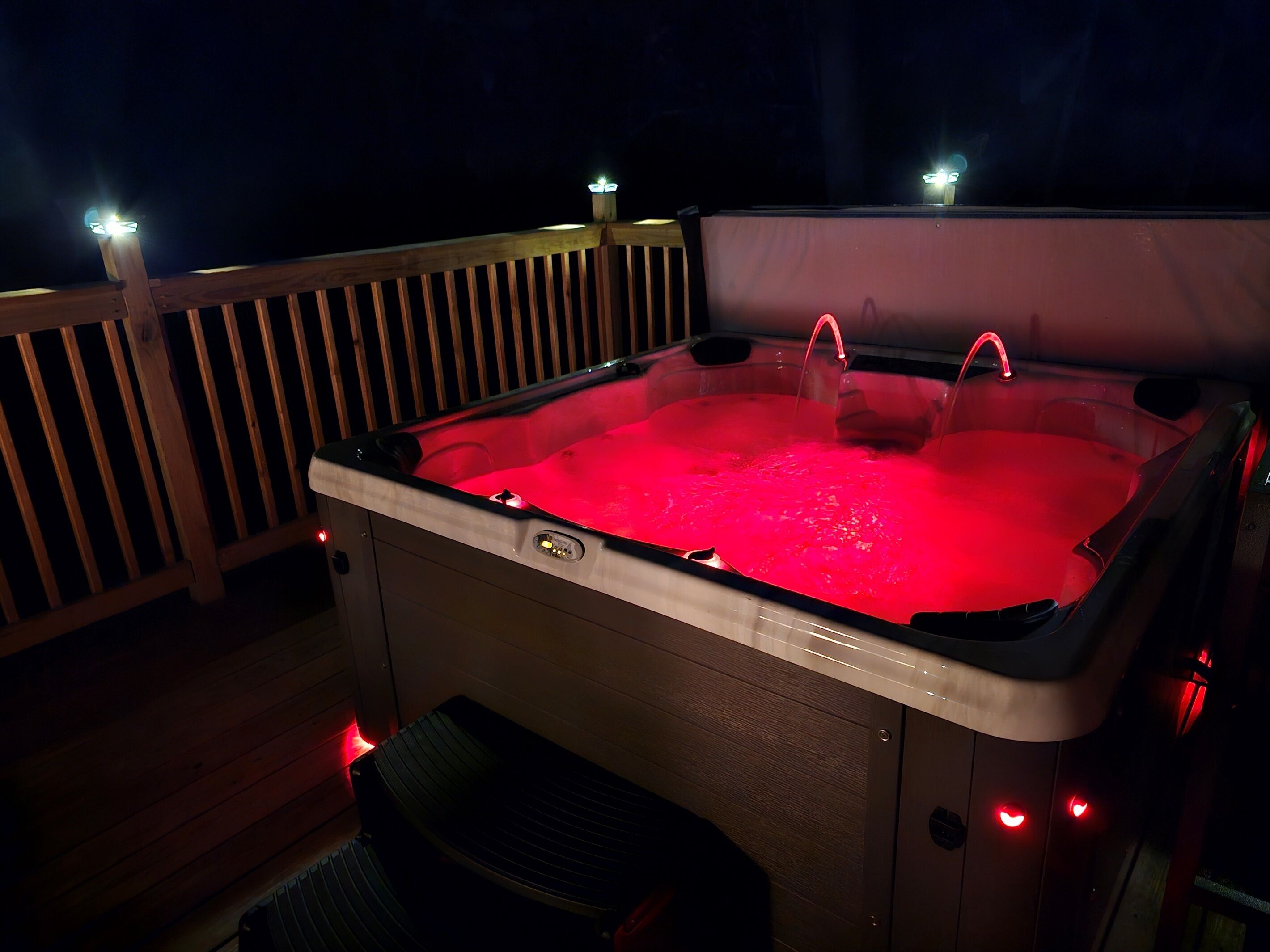 Un jacuzzi con terraza de madera y barandas, rodeado de iluminación nocturna.