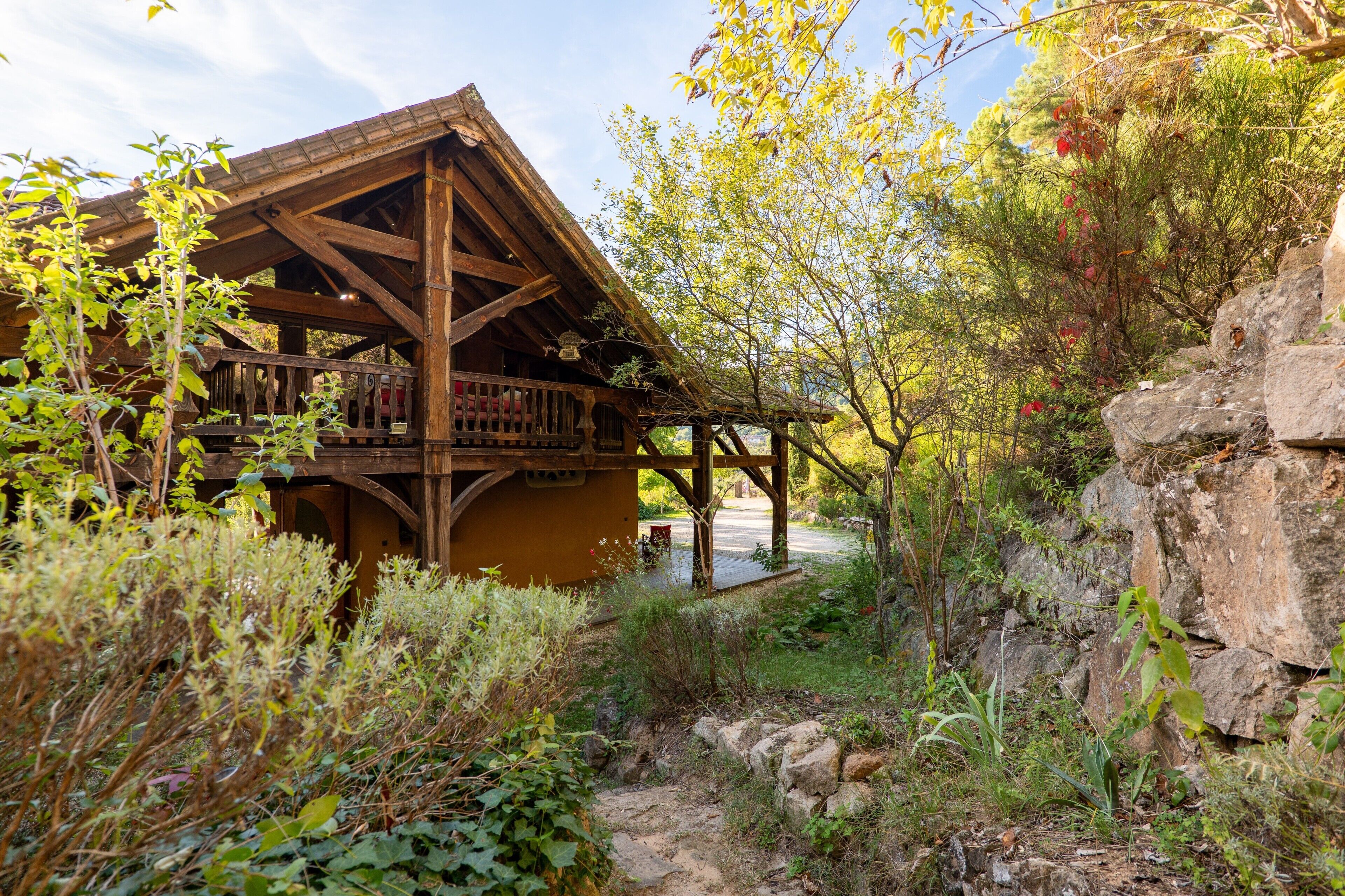 A rustic wooden cabin with a balcony, surrounded by natural vegetation and a rocky landscape.