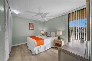 Een hotelkamer met een bed, een plafondventilator, een raam met jaloezieën en uitzicht op palmbomen.