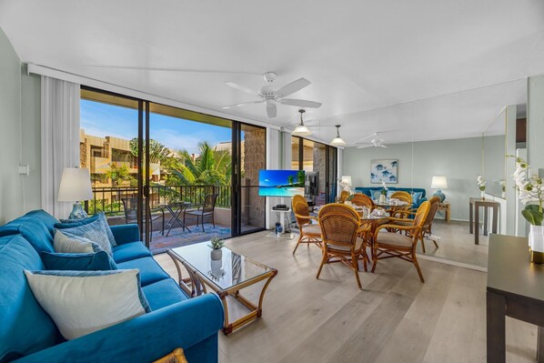 Een ruime woonkamer met een blauwe bank, een eettafel en stoelen, een televisie, en een balkon met uitzicht op palmbomen en gebouwen.