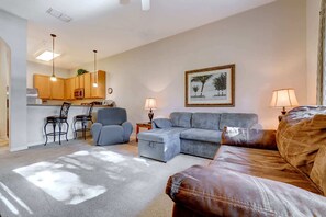 A living room with a sofa, armchair, and a small table with a lamp. There is a kitchen area with wooden cabinets and a dining table with chairs. - 3BR escape near Disney w/ balconies (Davenport)