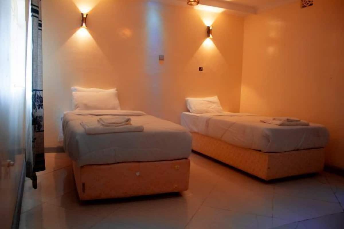 Una habitación con dos camas, cada una con ropa blanca y una toalla doblada. Hay luces fijadas a la pared en el techo.