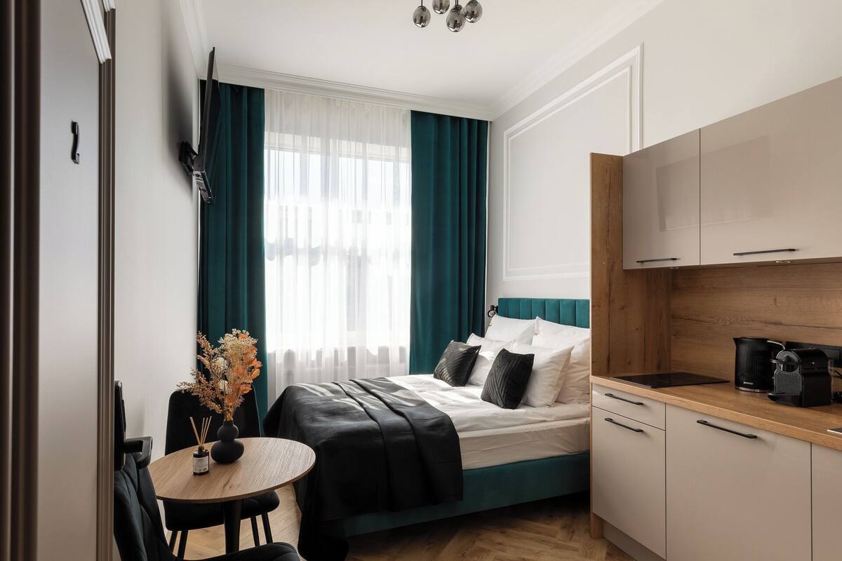 Una habitación de hotel compacta con una cama, una pequeña mesa de comedor, una silla, una kitchenette y un ventanal con cortinas.