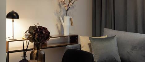 A modern living room with a grey sofa, a round coffee table, a framed cityscape picture, a vase with flowers, and two hanging pendant lights.