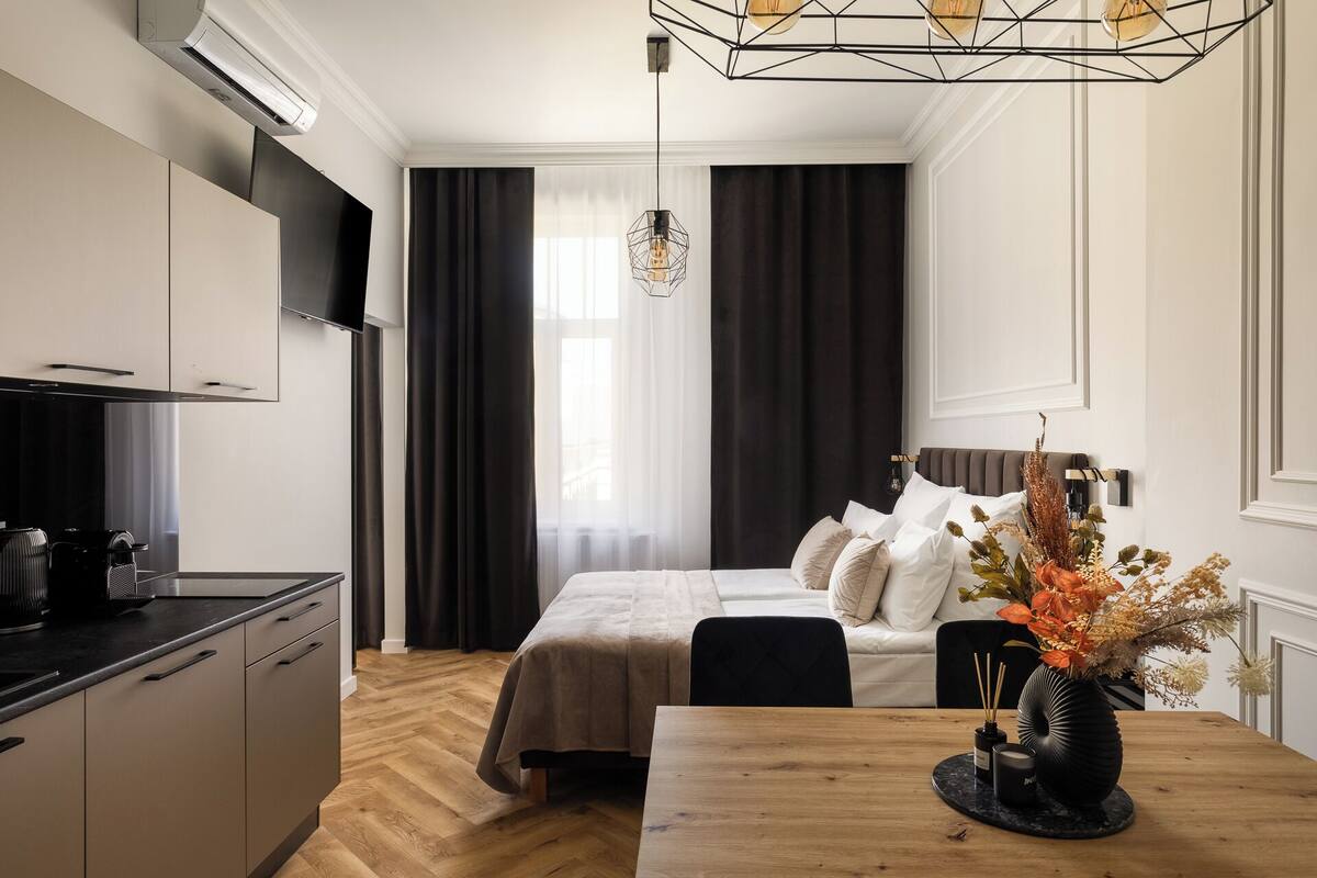 Una habitación de hotel moderna con kitchenette, una cama con cabecera, una mesa de comedor con un florero y un gran ventanal con cortinas.
