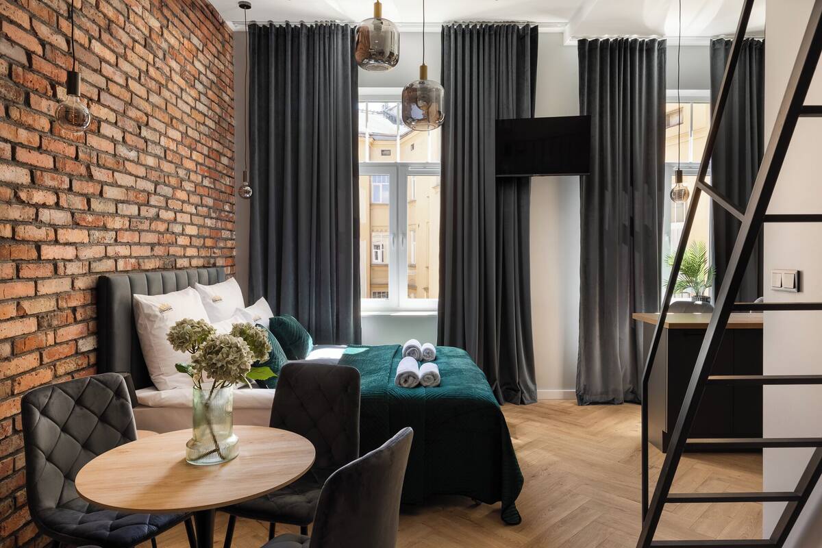 Habitación de hotel moderna con pared de ladrillo, sofá, cama con colcha verde, una mesa redonda de madera y vistas a edificios a través de amplias ventanas.