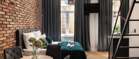 Habitación de hotel moderna con pared de ladrillo, sofá, cama con colcha verde, una mesa redonda de madera y vistas a edificios a través de amplias ventanas.