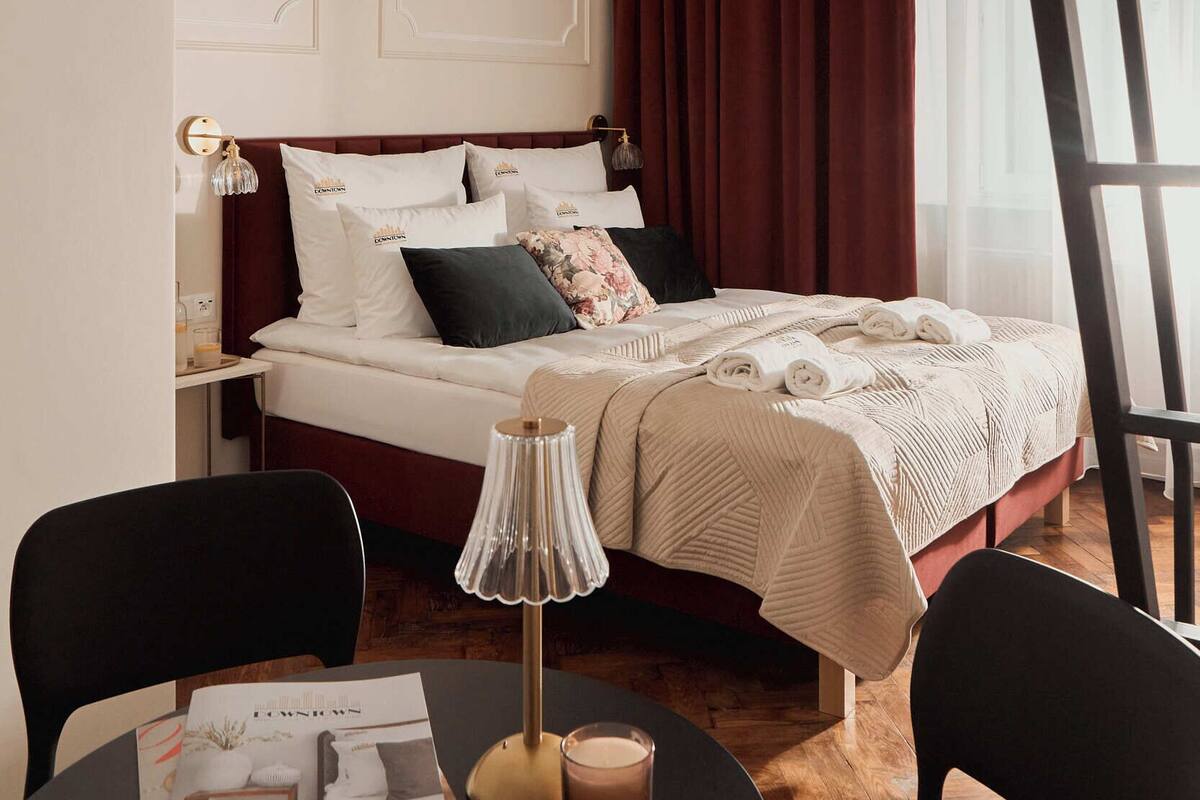 Una habitación de hotel con una cama, una mesita de noche, una lámpara, una revista sobre la mesa y una ventana con cortinas.
