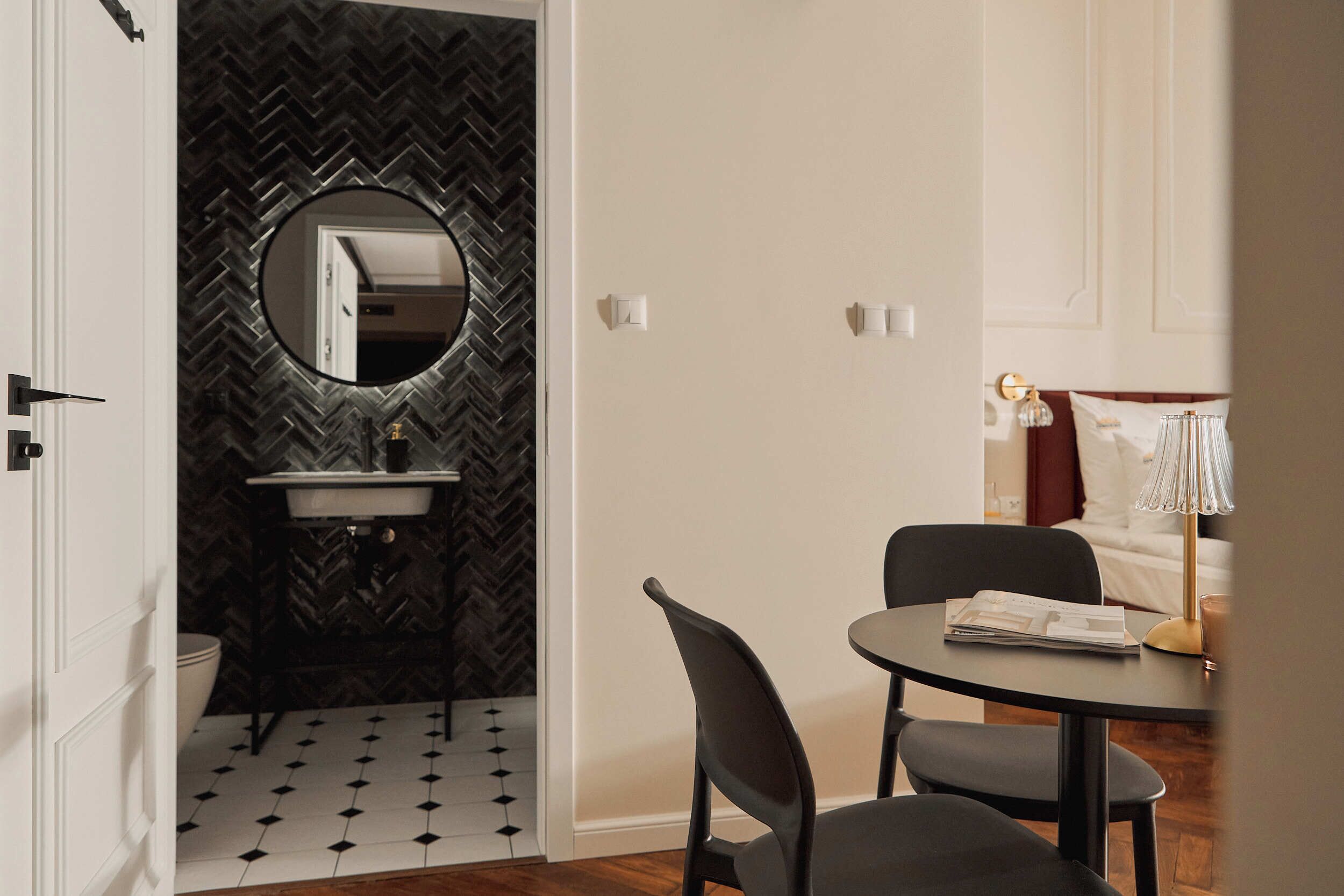 A compact kitchen with a herringbone-patterned backsplash, a round mirror, and a sink. There is a dining area with a table and chairs, and a bed with a lamp in the background.