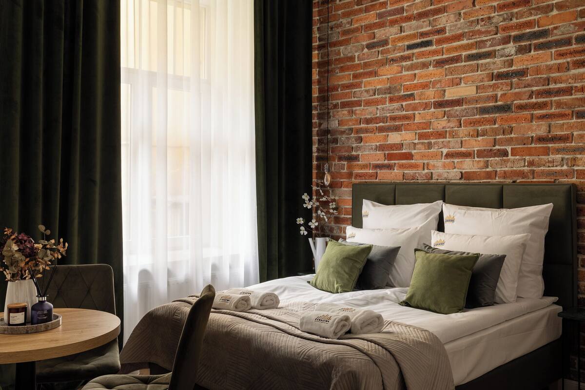 Habitación de hotel con pared de ladrillo, cama con almohadas verde y blanco, una mesa redonda con un florero y velas, y vistas al exterior a través de cortinas traslúcidas.
