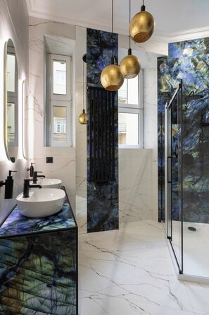 Ein modernes Badezimmer mit Marmorarbeitsplatten, zwei Waschbecken, goldenen Pendelleuchten und einer Duschkabine aus Glas.