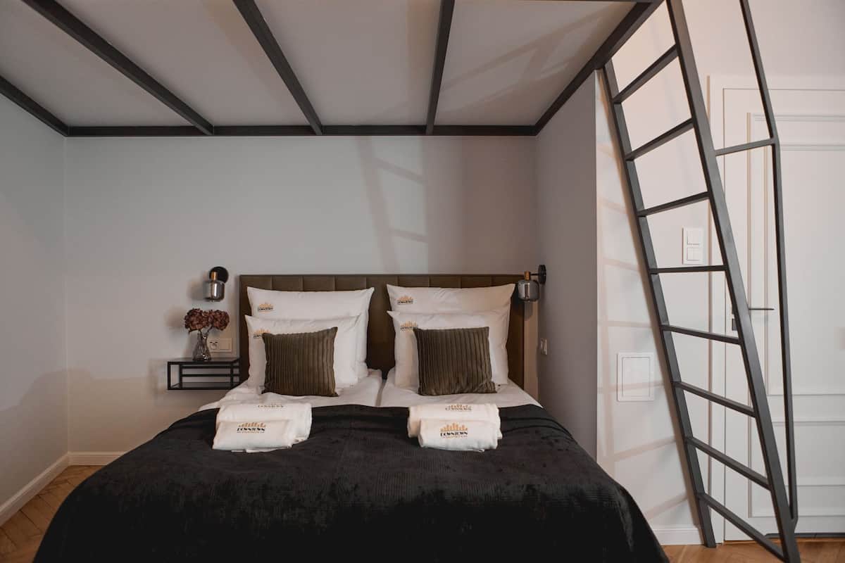 Una cama bien hecha con dos almohadas y dos toallas dobladas, una mesita de noche con un florero y una escalera apoyada en la pared.