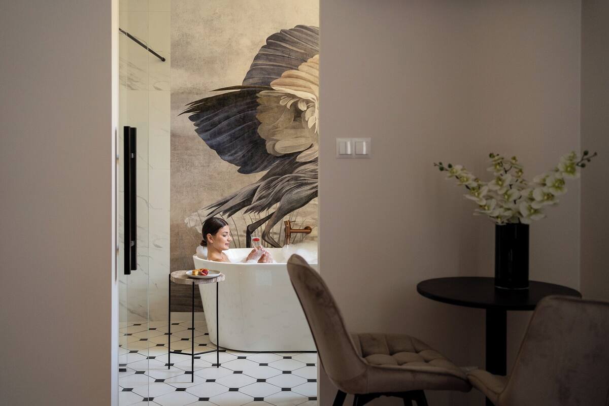 Un baño moderno con un gran mural de un pájaro, una bañera, una mesita redonda con una fuente de fruta y un florero con flores blancas.