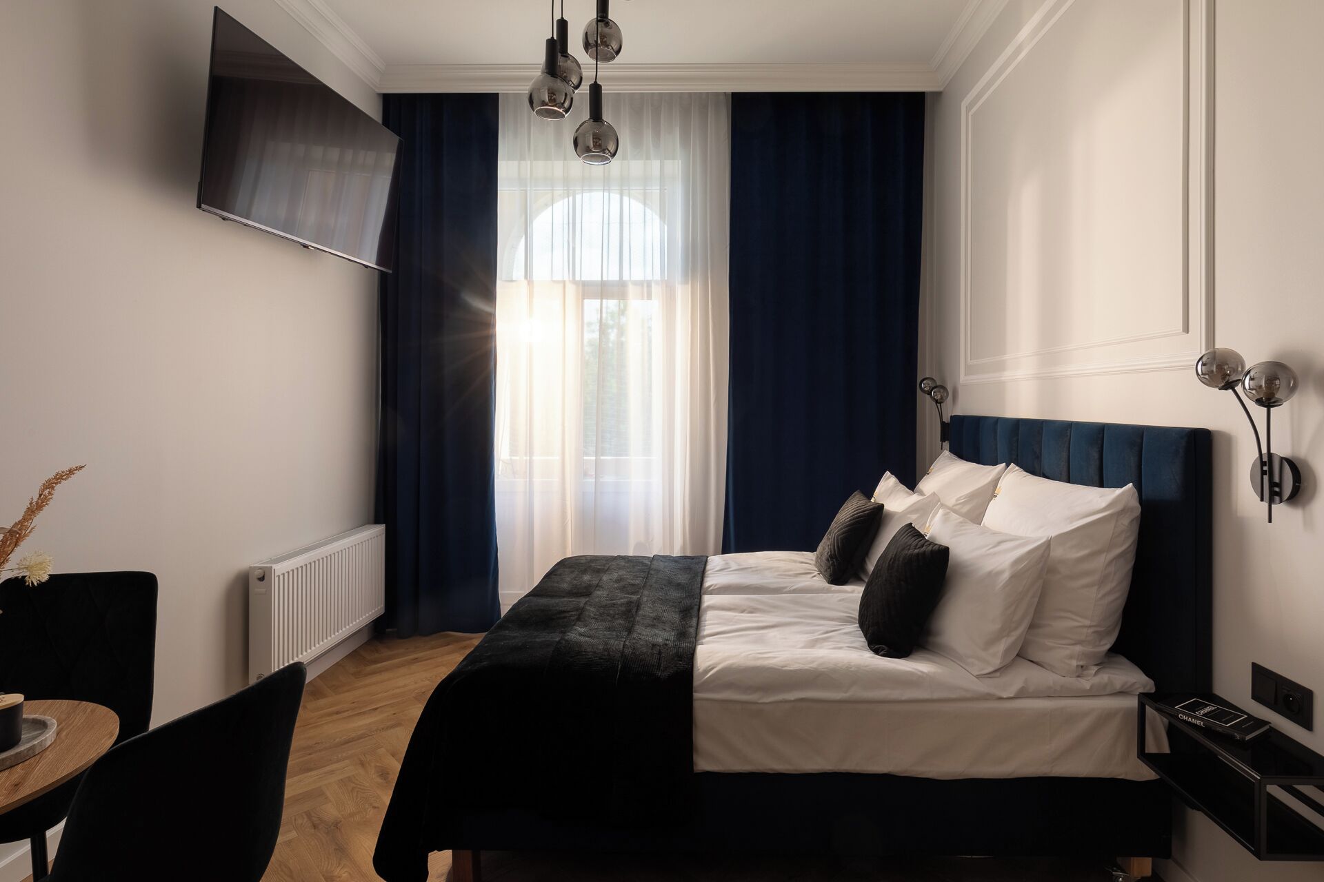 Et moderne hotelværelse med en stor seng, et vægmonteret fladskærms-tv, et spiseområde med bord og stole samt et vindue med gardiner.