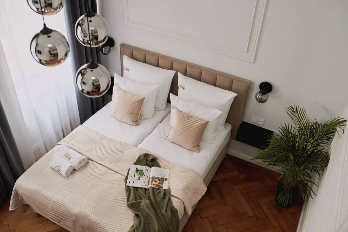 Una cama bien tendida con un cabecero color beige, almohadas blancas y una manta verde. A la derecha hay una planta en maceta y una lámpara de pared sobre la cama.