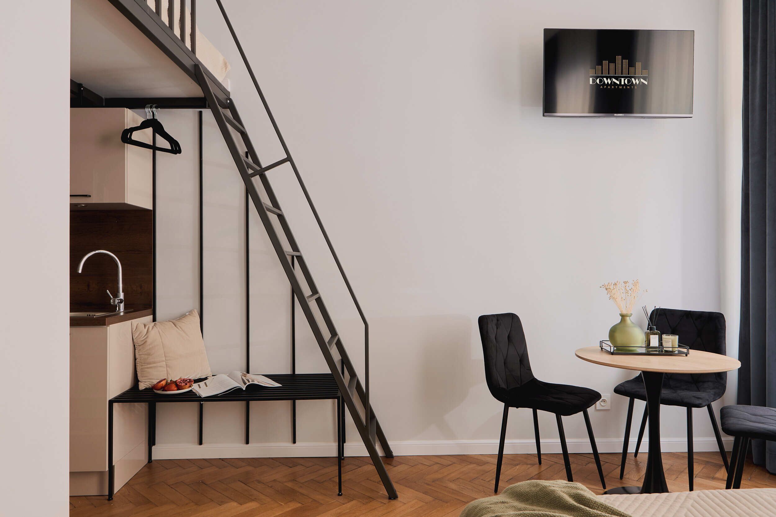 A modern apartment with a loft-style kitchen, a dining area with two chairs, a TV mounted on the wall, and a staircase.