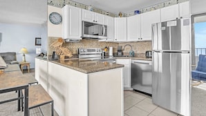 A compact kitchen with white cabinets, a stainless steel refrigerator, a microwave, and a coffee maker. - Cozy Oceanfront Condo with Heated Pools & Views (Carolina Beach)