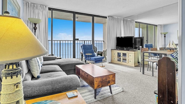 A modern hotel room with a balcony, a sofa, a coffee table, a TV, and a dining area. - Cozy Oceanfront Condo with Heated Pools & Views (Carolina Beach)
