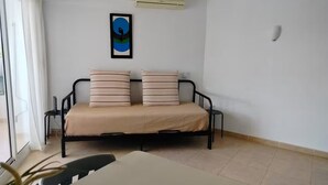 Un petit appartement d’une seule pièce, comprenant un lit, une table et une chaise. Il y a une fenêtre avec des rideaux, un climatiseur fixé au mur et un tableau encadré accroché au mur.
