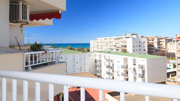 Un balcon avec vue sur la mer, des bâtiments blancs et un auvent rouge.