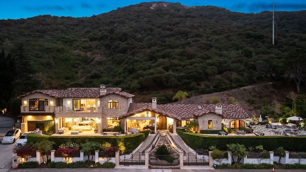 Una casa de dos pisos con un patio, un camino de entrada y montañas al fondo.