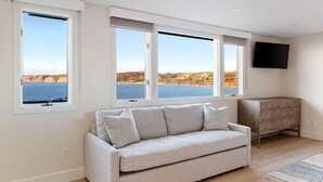 A living room with a sofa, a cabinet, and a TV mounted on the wall. There are three windows providing a view of the sea and cliffs. - New - Modern Studio - Stunning Ocean View (San Diego)