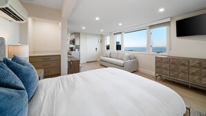A modern bedroom with a large bed, a sofa, and a TV. There is a kitchen area with cabinets and a microwave. The room has a large window with a view of the sea. - New - Modern Studio - Stunning Ocean View (San Diego)
