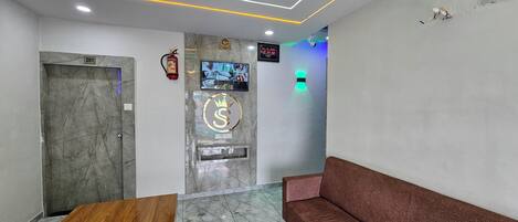 A hotel reception area with a marble reception desk, a sofa, a fire extinguisher, a digital clock, and a television displaying images.