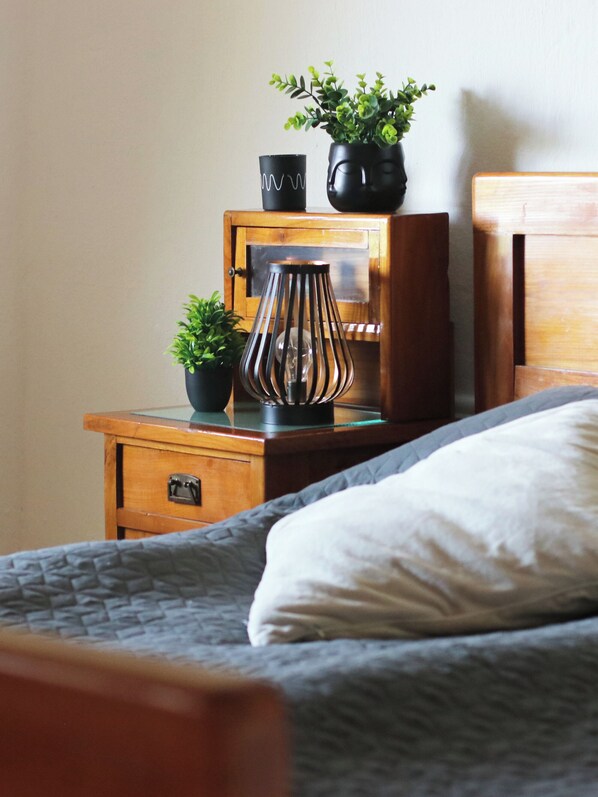 A neatly made bed with a white pillow and a gray blanket, a wooden nightstand with a small potted plant, a black vase, and a decorative metal lamp.