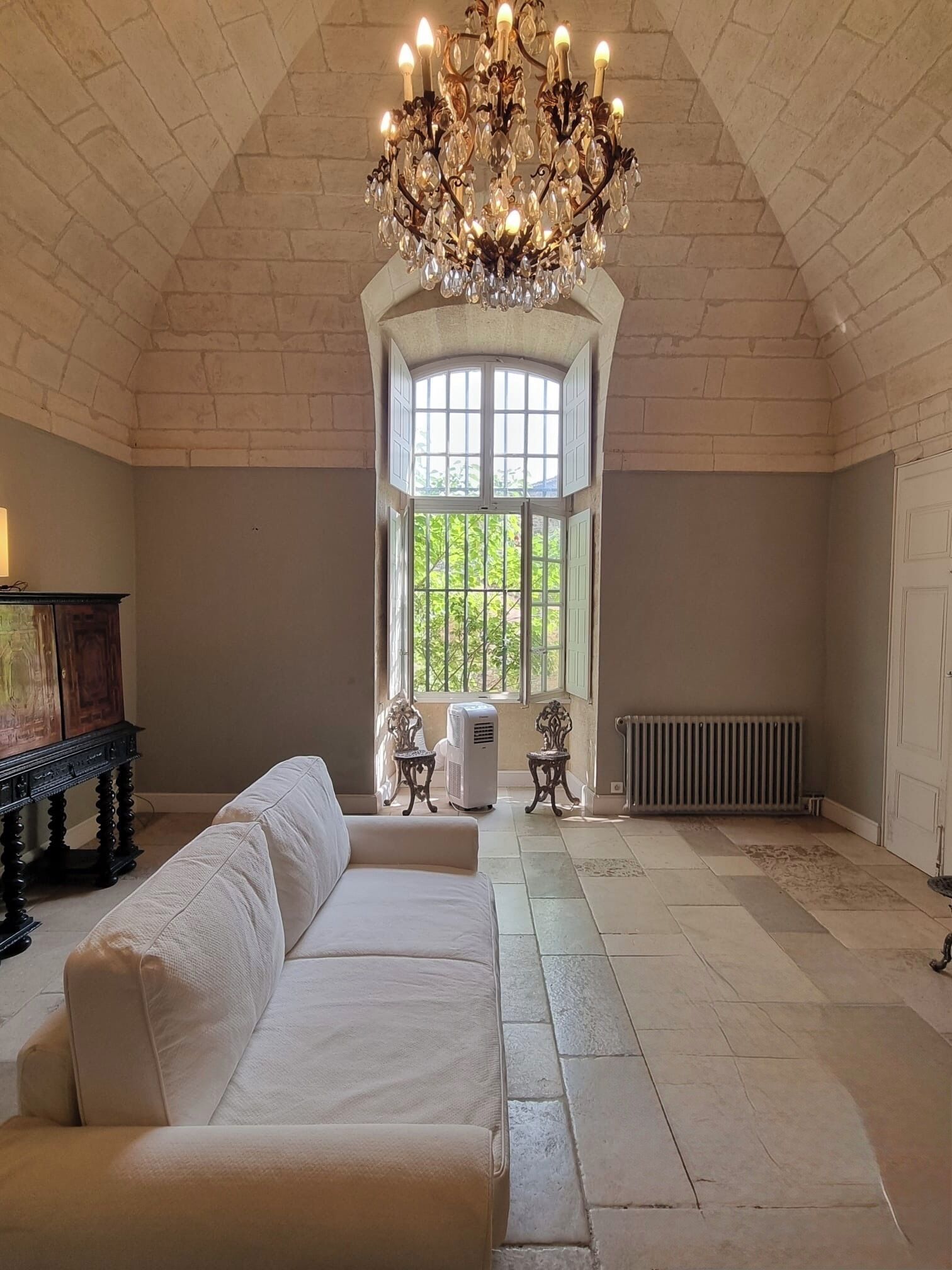 A spacious room with a high arched ceiling, a large window with a view of greenery, a white sofa, a chandelier, and a radiator.