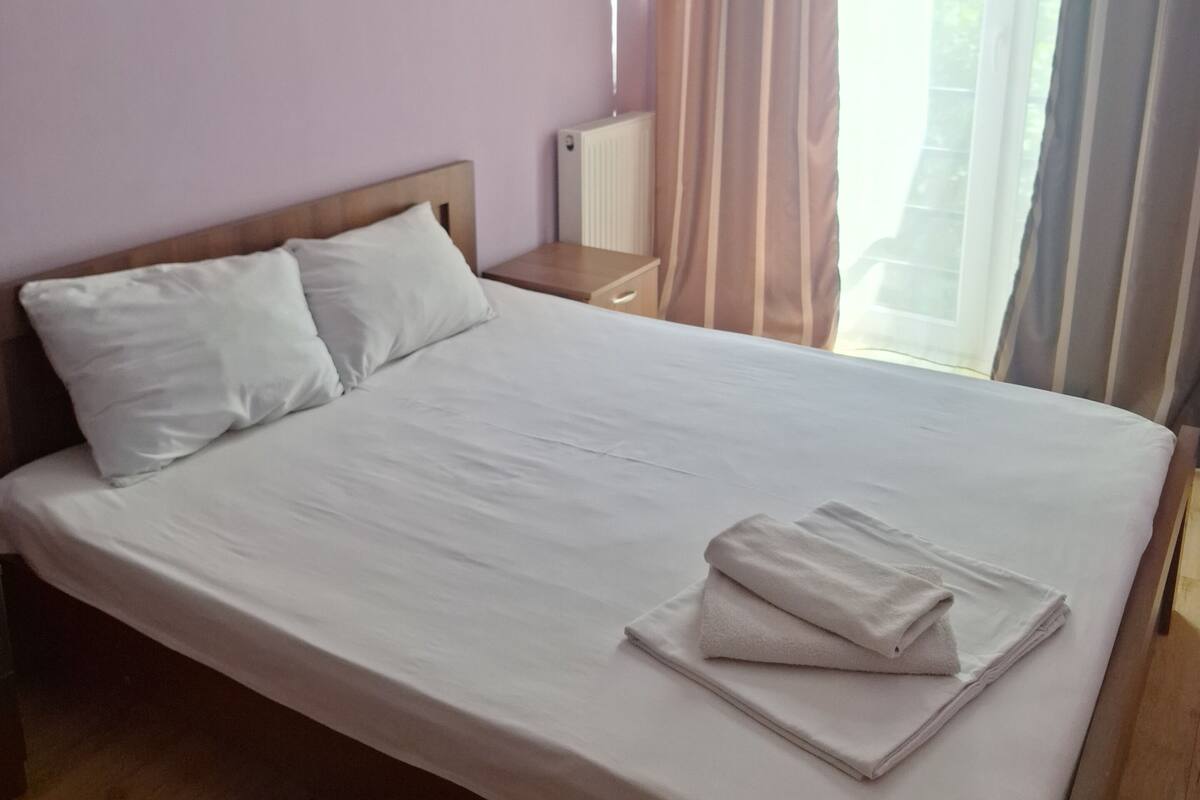 A hotel room with a bed, a nightstand, an air conditioning unit, and a window with curtains.