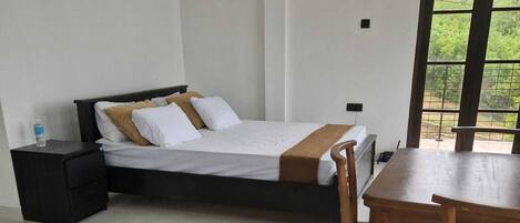 A hotel room with a bed, a desk, and a chair. There is a window with a view of greenery outside.