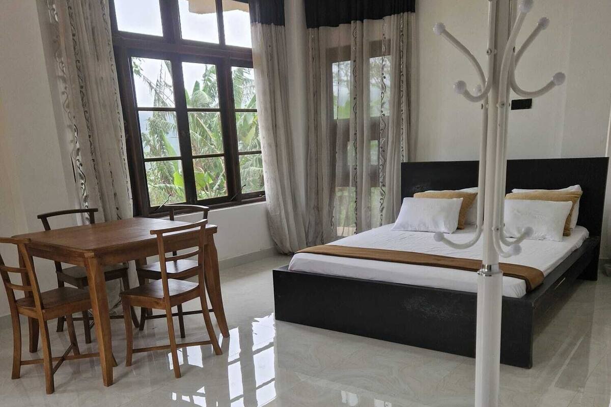 Una habitación de hotel con una cama, una mesa de comedor con sillas y un ventanal con cortinas.