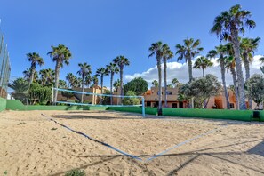 Un terrain de beach-volley sablonneux, avec un filet, entouré de palmiers et de bâtiments.