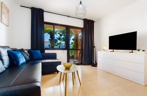 A modern living room with a black sofa, a round coffee table, a TV on a white cabinet, and a large window with curtains. - Coral Bliss Apartment with Shared Pool, Private Terrace, and Wi-Fi (Corralejo)