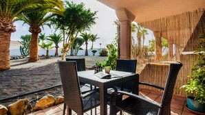 An outdoor patio with wicker furniture, a table with a book and a mug, and a view of palm trees and the sea. - Coral Bliss Apartment with Shared Pool, Private Terrace, and Wi-Fi (Corralejo)