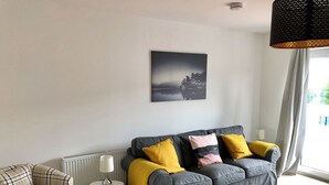 A living room with a grey sofa, yellow cushions, a plaid armchair, a small white side table, and a framed picture on the wall.