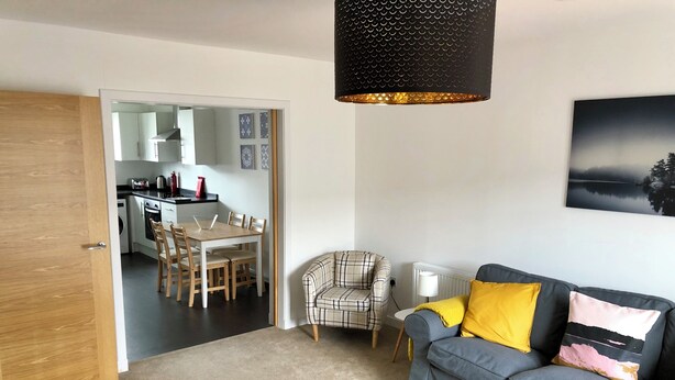 A living room with a grey sofa, a plaid armchair, a coffee table, and a kitchen in the background.