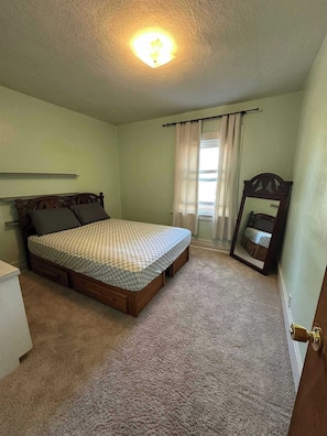 Quarto com uma cama de solteiro, mesa de cabeceira, cômoda, luminária no teto e janela com cortinas.