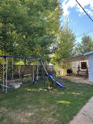 Un patio trasero con un tobogán, un conjunto de columpios y una zona de barbacoa.