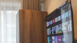 A hotel room with a wooden wardrobe, a cushioned chair, and a mounted TV displaying various streaming apps. - Columbus Hotel Eldoret (Eldoret)
