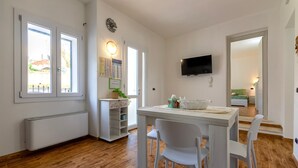 A modern living room with a dining table, white chairs, a TV mounted on the wall, and a view through the open door. - Casa Rinascita with Private Pool (Nulvi)